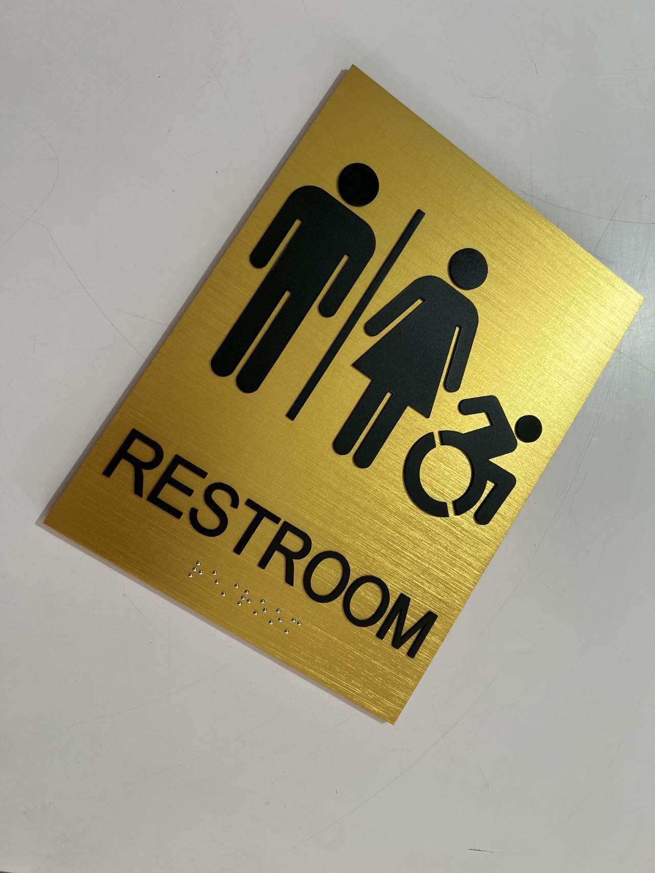 Angled view of a brushed gold accessible restroom sign with black pictograms and Braille